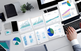 Person analysiert Datencharts und -grafiken auf Computerbildschirm, umgeben von Planungsmaterialien und modernem Arbeitsplatz.